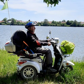 Claus sitzt auf seinem bepackten E-Dreirad am Donau-Ufer und schaut sich die Landschaft an.