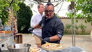 Nero ist ein etwa 50-jähriger Mann mit schwarzer Hornbrille. Er trägt ein kleines Mirko und schiebt gerade einen hölzernen Pizzaschieber unter eine frische Pizza.
