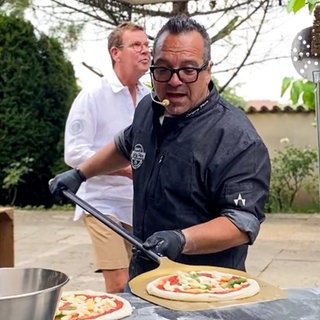 Nero ist ein etwa 50-jähriger Mann mit schwarzer Hornbrille. Er trägt ein kleines Mirko und schiebt gerade einen hölzernen Pizzaschieber unter eine frische Pizza.