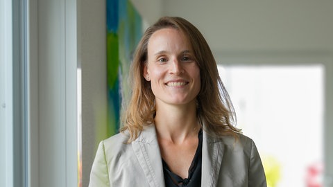 2015 erhält Katrin die Diagnose Endometriose. In ihrem Wohnzimmer steht sie im grauen Blazer und blickt mit einem freundlichen Lächeln direkt in die Kamera.