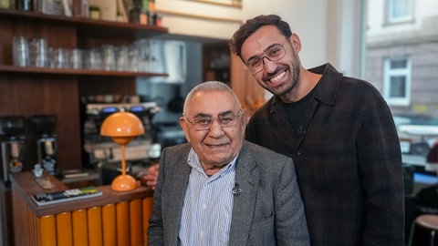 Barış und sein Opa Cemal stehen vor der Bar im Café Kalo. Barış hat ein Arm über die Schulter seines Opas gelegt und sie lachen in die Kamera.