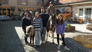 Das Hospiz in Dudenhofen bekommt regelmäßig Besuch von zwei Pferden. Auch Tiere, wie Eulen und Alpakas waren schon auf dem Hof. Christian lächelt gemeinsam mit drei Kindern, den zwei Pferden und der Pferdebesitzerin in die Kamera.