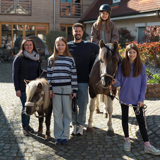 Das Hospiz in Dudenhofen bekommt regelmäßig Besuch von zwei Pferden. Auch Tiere, wie Eulen und Alpakas waren schon auf dem Hof. Christian lächelt gemeinsam mit drei Kindern, den zwei Pferden und der Pferdebesitzerin in die Kamera.