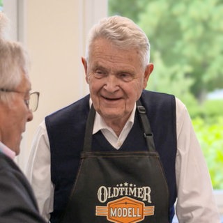Dietmar ist im Gespräch mit zwei anderen Teilnehmern des Kochkurses für Herren über 70. Er hat weiße Haare, trägt einen dunklen Pullunder und eine schwarze Kochschürze, auf der „Oldtimer Modell“ steht. 