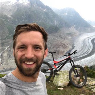 Junger Mann mit braunen Haaren lächelt in die Kamera, im Hintergrund Berge und ein Fahrrad