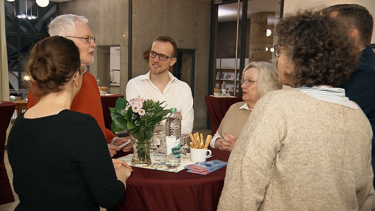 Gruppe von Menschen steht um einen Tisch und unterhält sich.