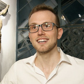 Junger Mann mit Brille steht in einer Kirche vor einem Holzkreuz.