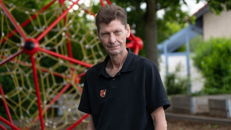Mann mit schwarzem T-Shirt vor einem Klettergerüst auf einem Schulhof.