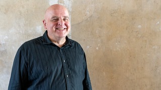 Mann mit Glatze steht vor einer Wand und lächelt