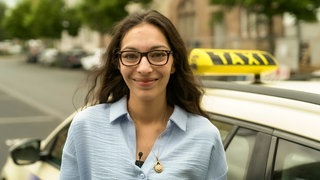 Junge Frau, mit braunen Haaren, dunkler Brille und blauer Bluse vor einem Taxi, dass an einer Straße steht.