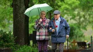 Alte Frau und Mann mit Regenschirm vor Bäumen.