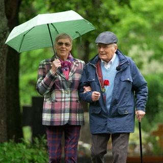 Alte Frau und Mann mit Regenschirm vor Bäumen.