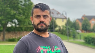 Ein Mann mit grauem Shirt steht auf einer Wiese und schaut traurig in die Kamera