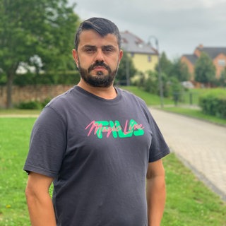 Ein Mann mit grauem Shirt steht auf einer Wiese und schaut traurig in die Kamera