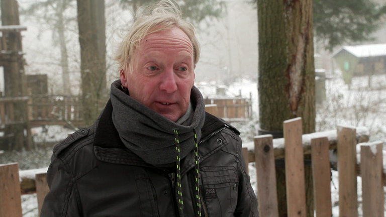 Ein älterer Mann steht mit weißen langen Haaren und dunkler Jacke auf einem Plateau des Baumhauses.
