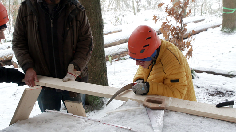 Ein Junge in gelber Jacke und orangenen Helm sägt einen Balken in zwei Teile.