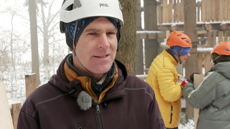 Ein Mann mit brauner Jacke, weißem Kletterhelm und Mütze, steht vor ein paar Kindern, die etwas am Baumhaus arbeiten.