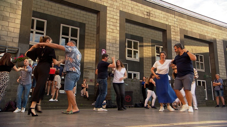 Mehrere Menschen tanzen gemeinsam auf einem Platz vor einem Haus.