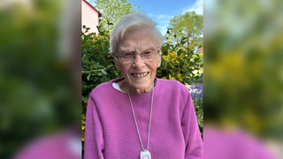 Alte Frau mit weißen Haaren und lila Pullover lächelt in Kamera.