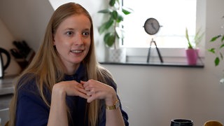 Eine junge Frau mit langen Haaren sitzt in einer Wohnung und lächelt an der Kamera vorbei.