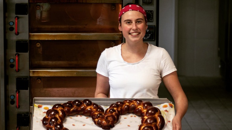 Junge Frau, in weißer Bäckerklufft, vor Ofen hält ein großes Hefezopfherz in die Kamera. 