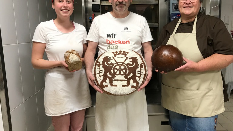 Drei Menschen von denen alle ein Brot in der Hand halten. Links eine junge Frau, in der Mitte ein älterer Mann und recht eine ältere Frau mit Schürze. Sie stehen vor einem Industrieofen. 