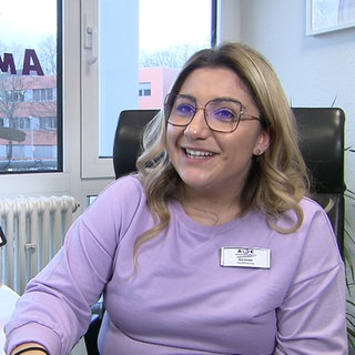 Junge Frau mit lila Pullover und eckiger Brille schaut an der Kamera vorbei. 