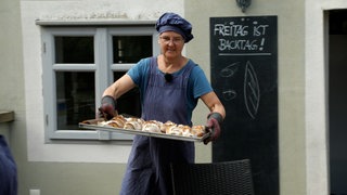 Frau mit blauer Mütze und Schürze trägt Blech mit Brot.