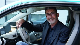 Gut gekleideter Mann auf dem Fahrersizt seines E-Autos durch die offene Tür schauend
