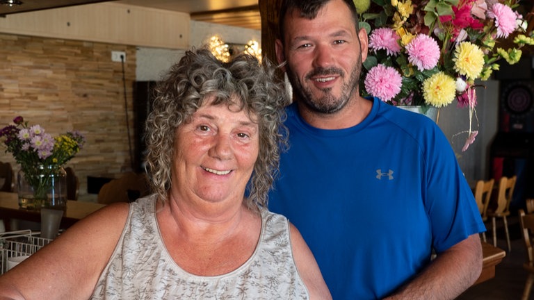Eine Frau mit grauen Locken steht neben ihrem Sohn mit blauem T-Shirt und schwarten Haaren vor einem Tresen. 