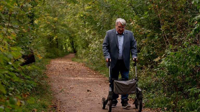 Ein Mann mit weißen Haaren spaziert mit Rollator durch den Wald.