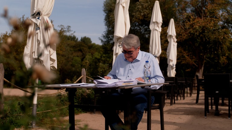 Mann im blauen Hemd sitzt an Tisch vor Sonneschirmen.