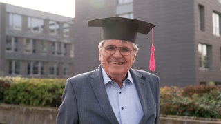 Ein älterer Mann mit Doktohut lächelt in die Kamera.