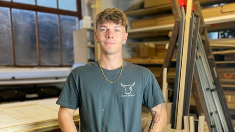 Junger Mann mit blauem T-Shirt und Arbeitshose in einer Schreinerei.