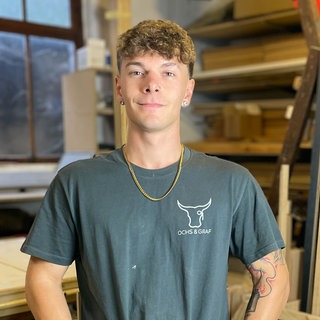 Junger Mann mit blauem T-Shirt und Arbeitshose in einer Schreinerei.