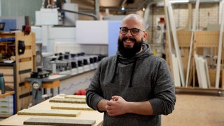 Junger Mann mit Glatze, Vollbart und schwarzen Kapuzenpullover steht in einer Schreinerei. 