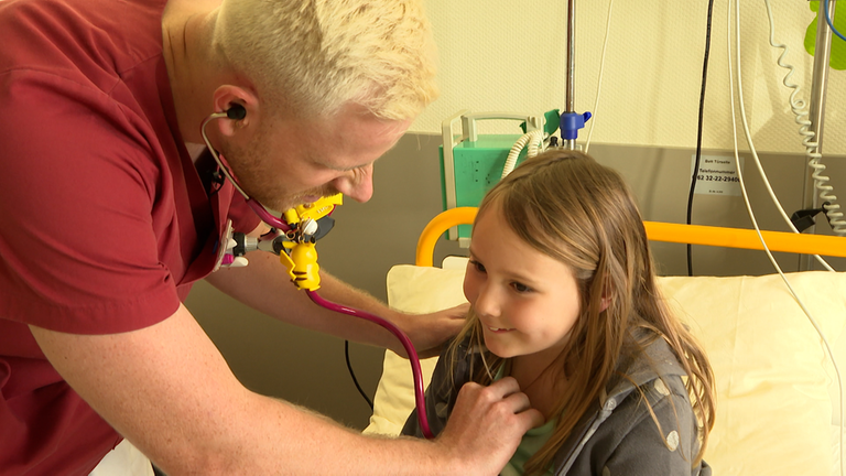 Aaron behandelt ein junges Mädchen im Krankenhauszimmer. Er hört die siebenjährige Patientin mit dem Stethoskop ab. 