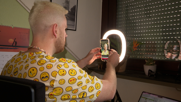 Aaron filmt sich selbst und nimmt mit dem Handy ein Video an seinem Schreibtisch in seinem Arztkittel auf. 