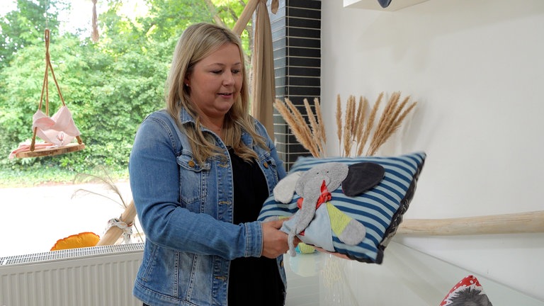 Frau mit langen blonden Haaren, einer blauen Jeansjacke und einem schwarzen Shirt darunter hält ein Kissen in ihren Händen. Das Kissen ist hell- und dunkelblau gestreift. Auf der Mitte des Kissens ist ein hellgrauer Elefant aufgenäht. Die Frau schaut auf das Kissen, welches sie in der Hand hält.