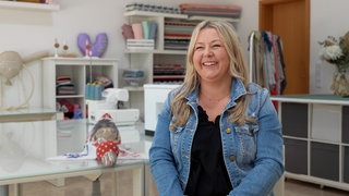 Frau mit langen blonden Haaren, einer blauen Jeansjacke und einem schwarzen Shirt darunter sitzt auf einem Stuhl und strahlt in die Kamera. Links neben ihr liegt ein Stofftier in Form einer Wildente. Im Hintergrund ist ihr Nähgeschäft zu sehen.