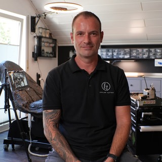 Mann in schwarzem T-Shirt schaut in Kamera, hinter ihm steht eine Liege zum Tätowieren.