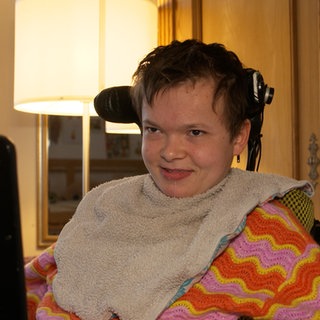 Junge Frau mit kurzen Haaren sitzt in einem Rollstuhl und lächelt