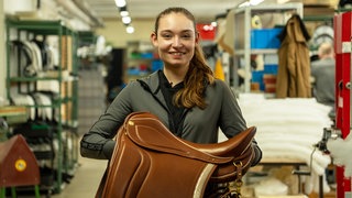 Junge Frau mit Sattel in der Hand steht lachend in einer Halle.