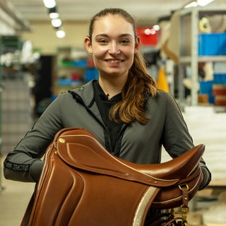 Junge Frau mit Sattel in der Hand steht lachend in einer Halle.