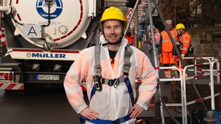 Junger Mann Anfang 20, trägt einen weißen Anzug, Sicherheitsgurte und einen gelben Helm. Im Hintergrund sieht man ein Lastwagen und Mitarbeiter der Kanalfirma.