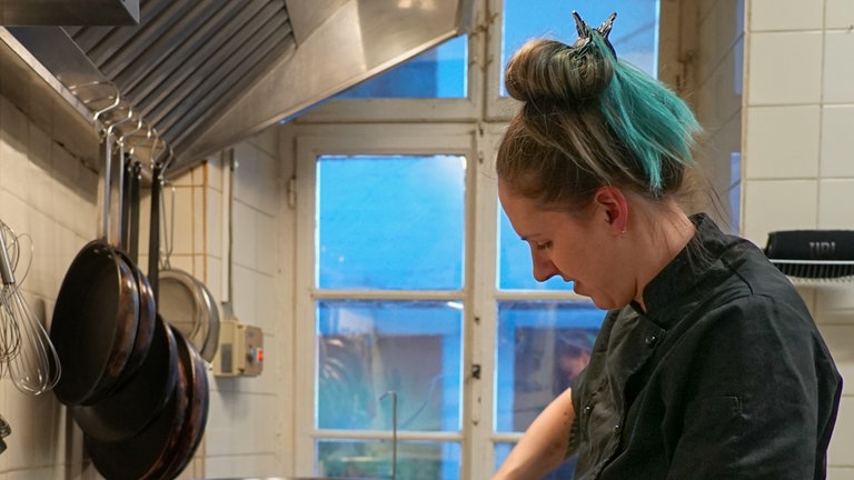 Junge Frau in schwarz, mit grüner Schürze steht am Herd und macht Dampfnudeln.