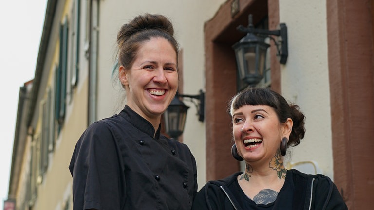 Zwei junge Frauen in schwarz gekleidet mit grüner Schürze stehen vor dem Eingang eines Lokals