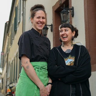 Zwei junge Frauen in schwarz gekleidet mit grüner Schürze stehen vor dem Eingang eines Lokals