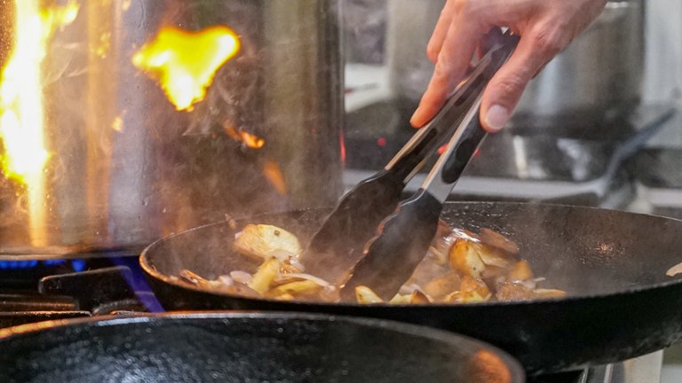 Pfanne mit Bratkartoffeln, die mit einer Grillzange gewendet werden.