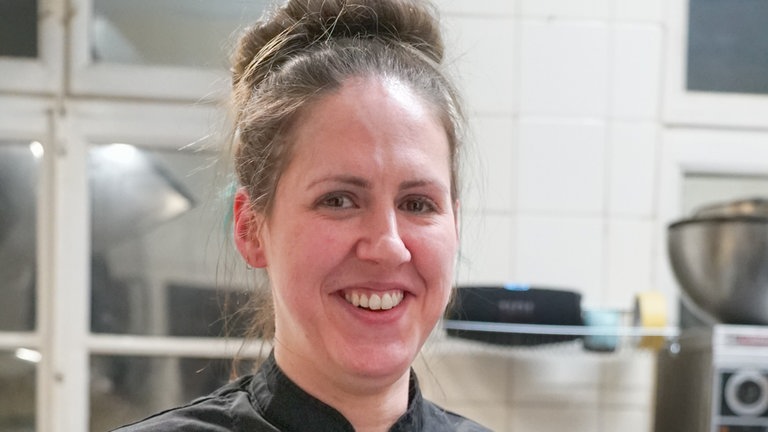 Junge Frau mit schwarzer Kochkleidung hält einen Teller mit Wurstsalat und Bratkartoffeln in der Hand.